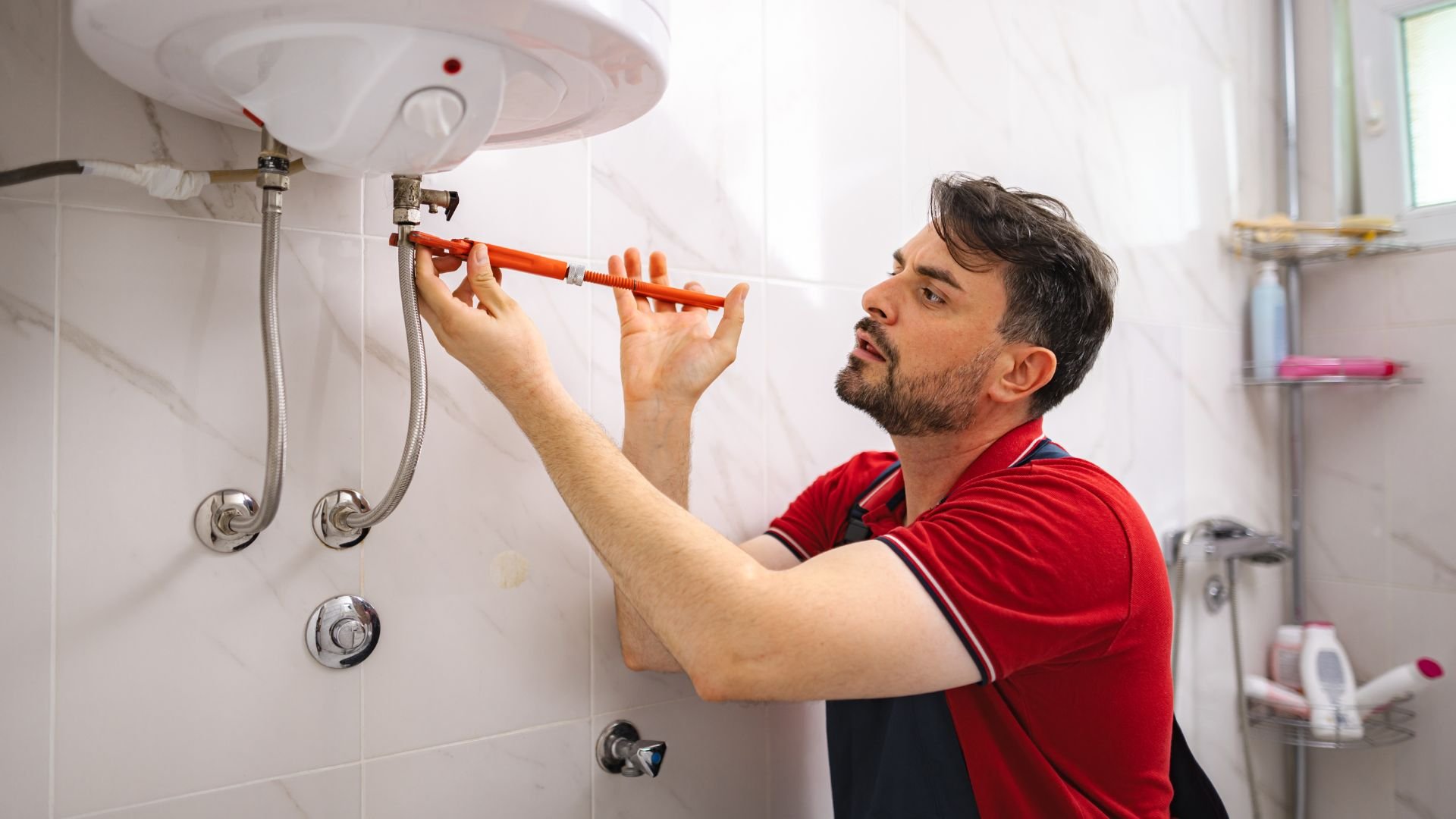 Plumber in red shirt fixing water heater pipes with wrench in bathroom